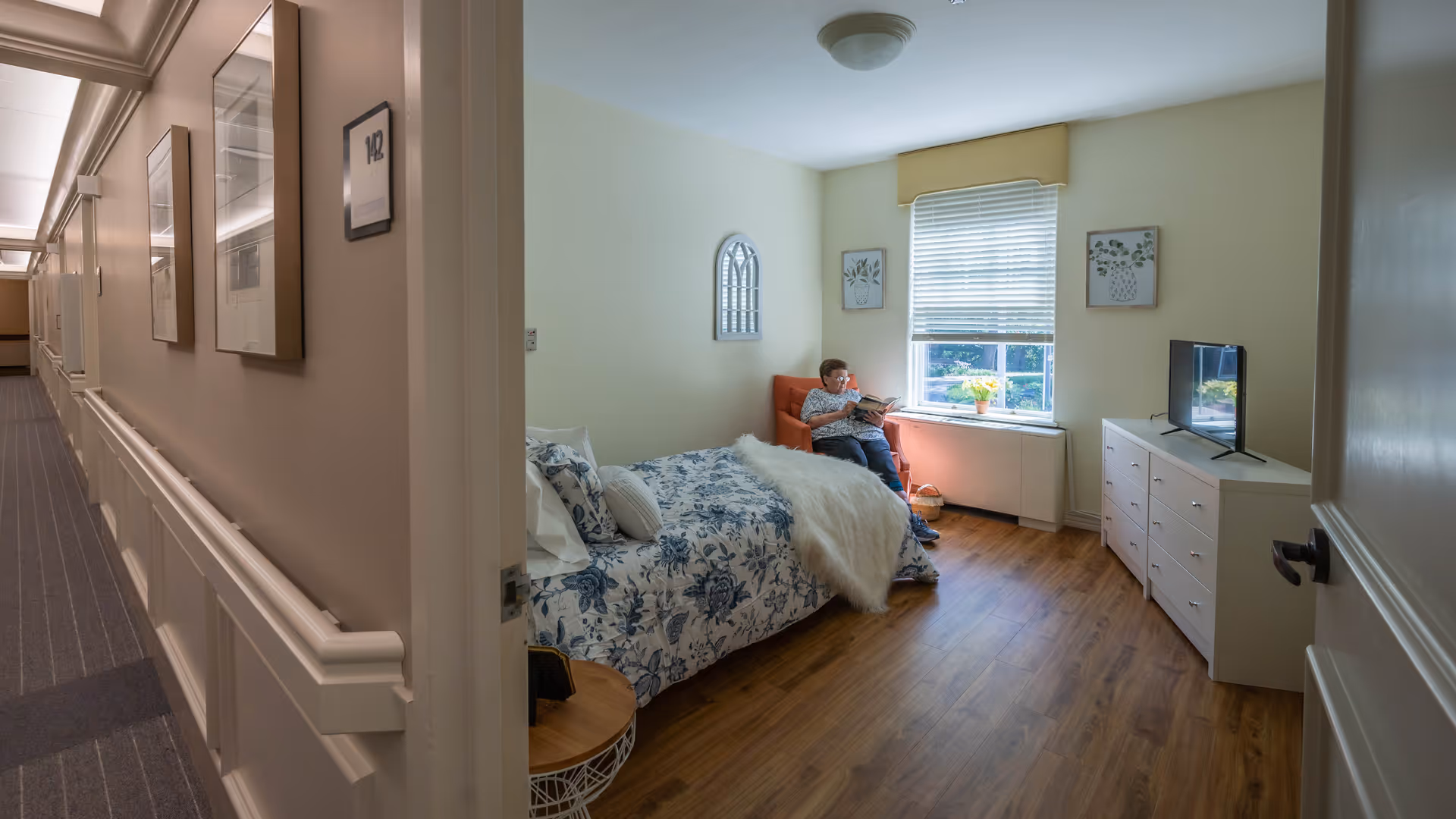 View of a senior living facility bedroom with a bed covered in a blue and white floral comforter, a small round side table, a dresser with a TV on top, and a window with blinds. A person is sitting in an orange armchair near the window reading a book. The room has light-colored walls and wood flooring. The image also shows part of the hallway outside the room.