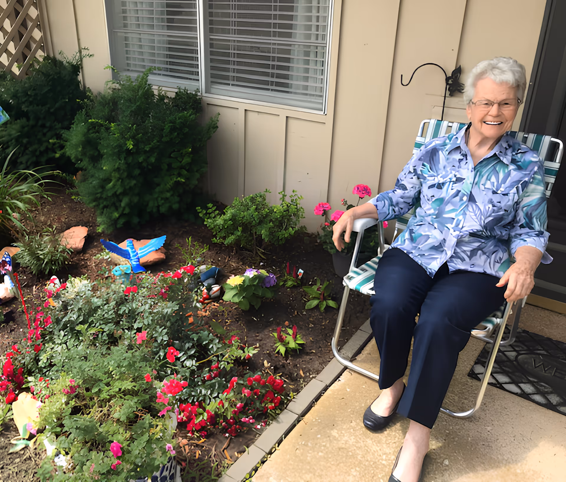 An elderly woman smiling while sitting on a patio chair beside a small flower garden outside a building.