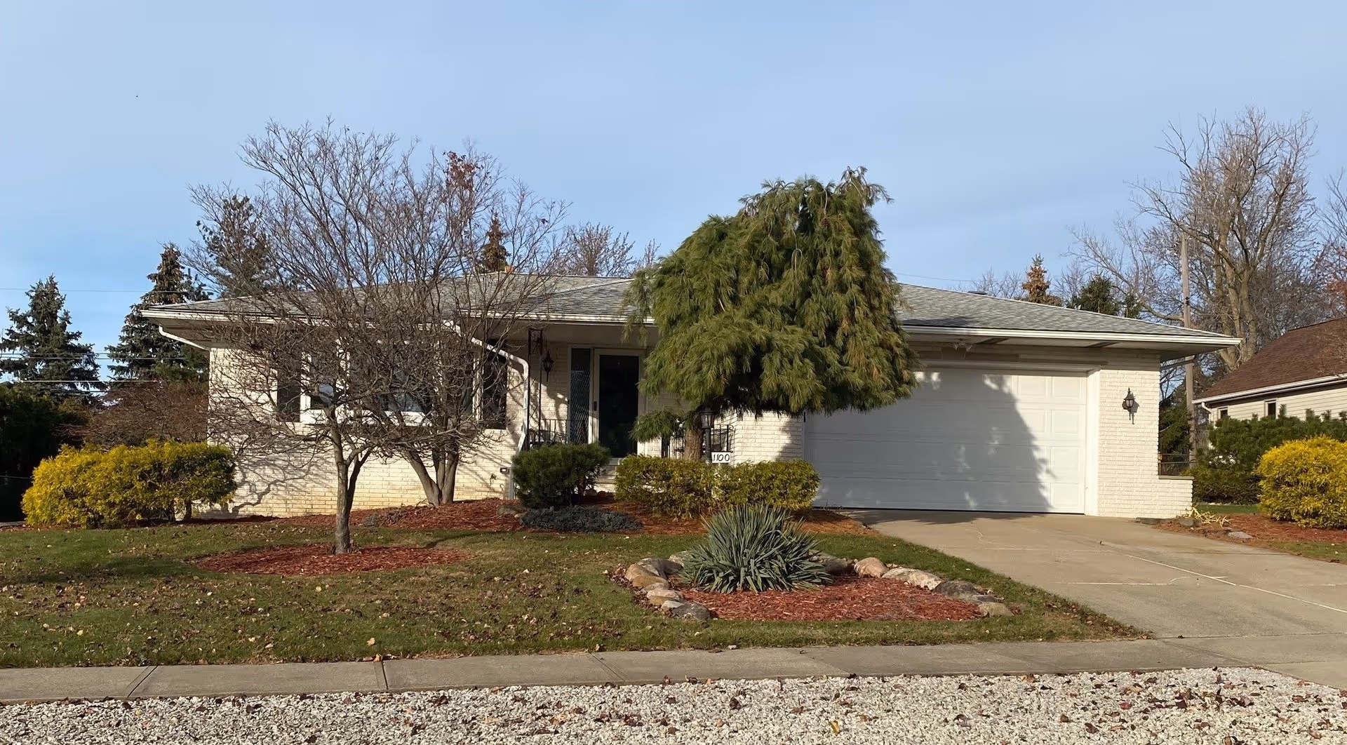 Exterior front view of a single-story residential building with a light-colored brick facade, a two-car garage, a driveway, and a landscaped front yard featuring trees, shrubs, and mulch beds.