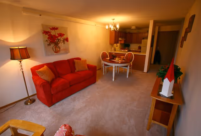 Interior view of a senior living facility apartment featuring a red sofa with two cushions, a floor lamp, a round dining table with two chairs, and a kitchen area with wooden cabinets and white appliances in the background. The room is carpeted and decorated with a floral painting and a small table with a plant and decorative item.