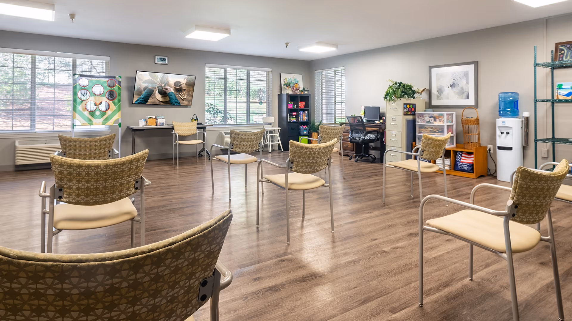 A spacious room with several chairs arranged in a semi-circle facing a wall-mounted TV. The room has large windows letting in natural light, a water cooler, a filing cabinet, a desk with a computer, and shelves with various items. The floor is wooden, and the walls are light-colored.