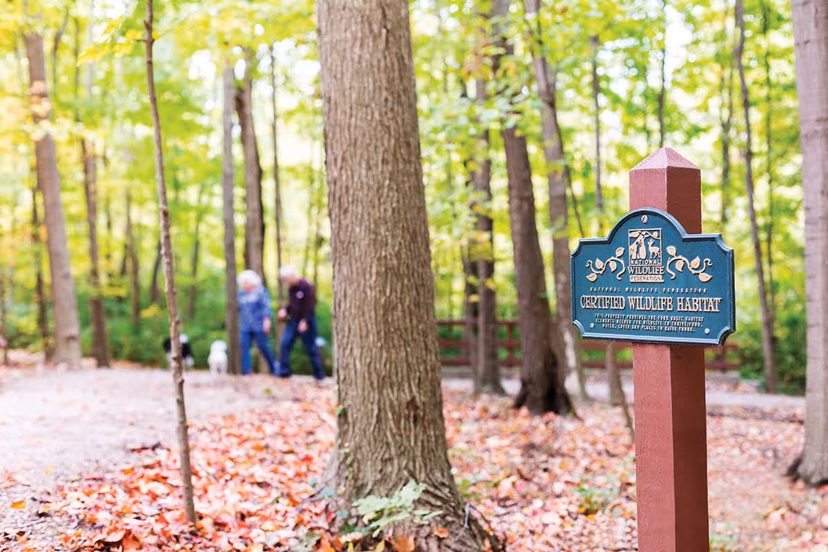 A wooded outdoor area with a signpost reading 'Certified Wildlife Habitat' in the foreground. In the background, two elderly people walk along a path surrounded by trees with green and autumn-colored leaves.