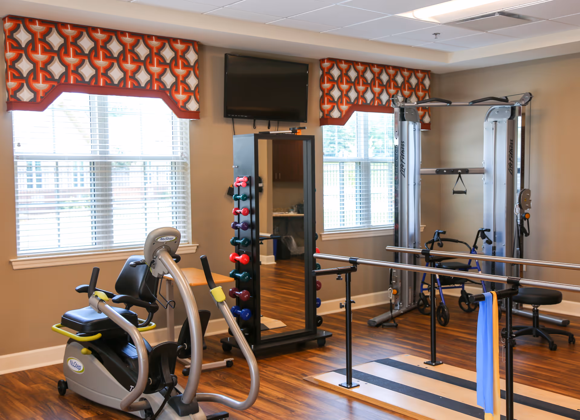 Bright interior exercise and physical therapy room with a recumbent bike, parallel bars, dumbbell rack, cable machine and wall-mounted TV.