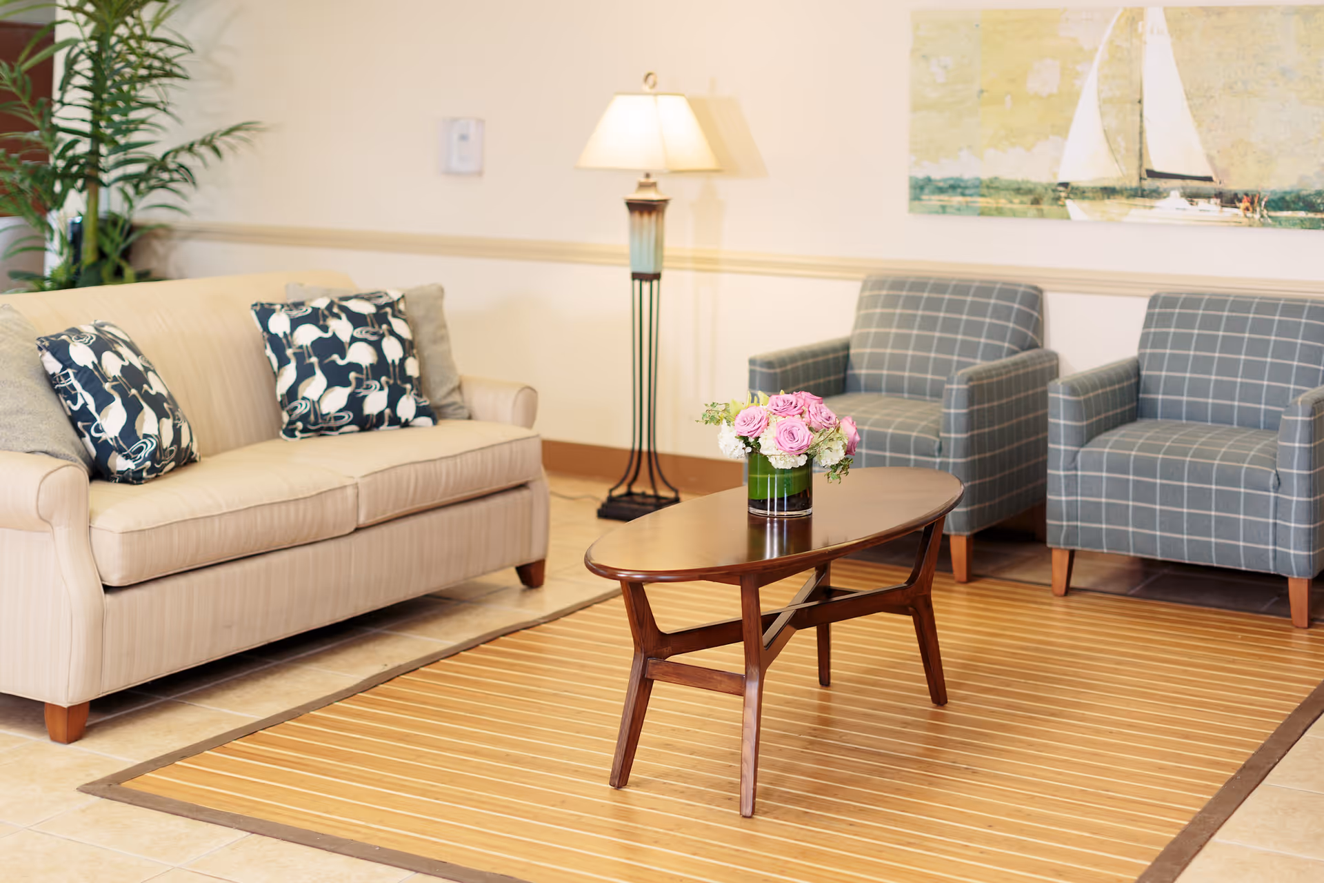 A bright sitting area with a beige sofa, two patterned armchairs, a wooden coffee table holding a vase of flowers, and a floor lamp.