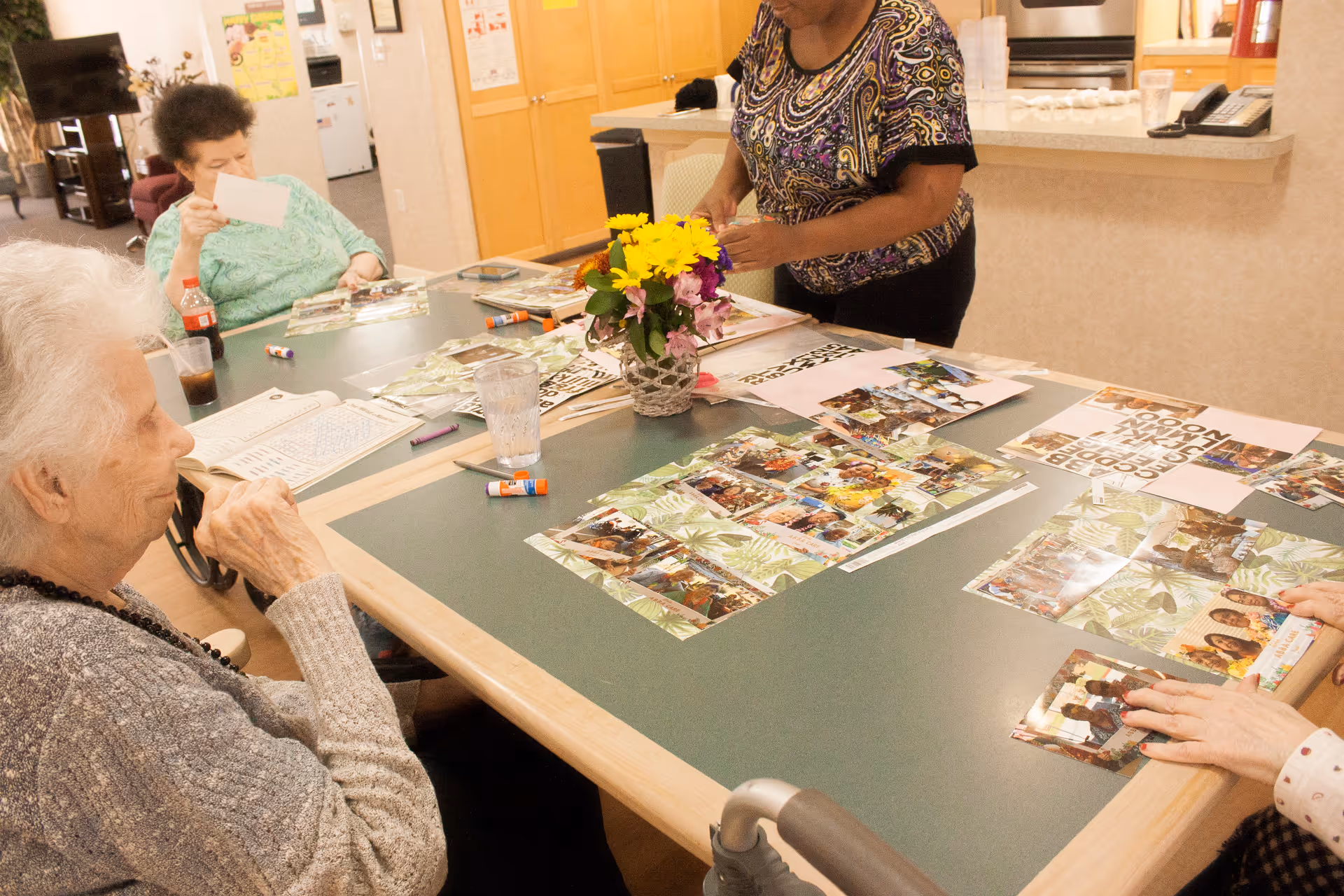 Several elderly women sitting and standing around a large table engaged in a craft activity involving photos and glue sticks in a well-lit room with a kitchen area in the background. A vase with yellow and purple flowers is placed on the table.