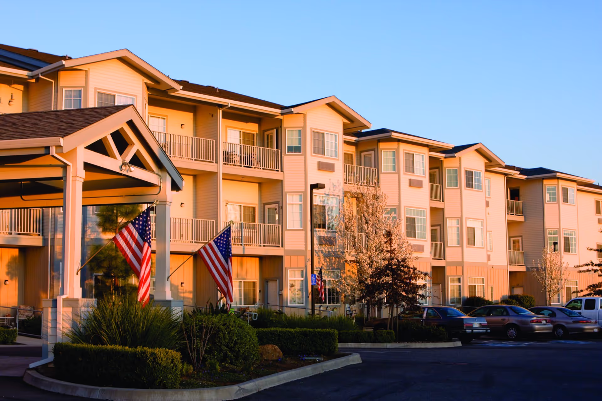 Exterior view of a multi-story senior living facility building with balconies, American flags, landscaped bushes, and a parking lot with several cars during sunset.