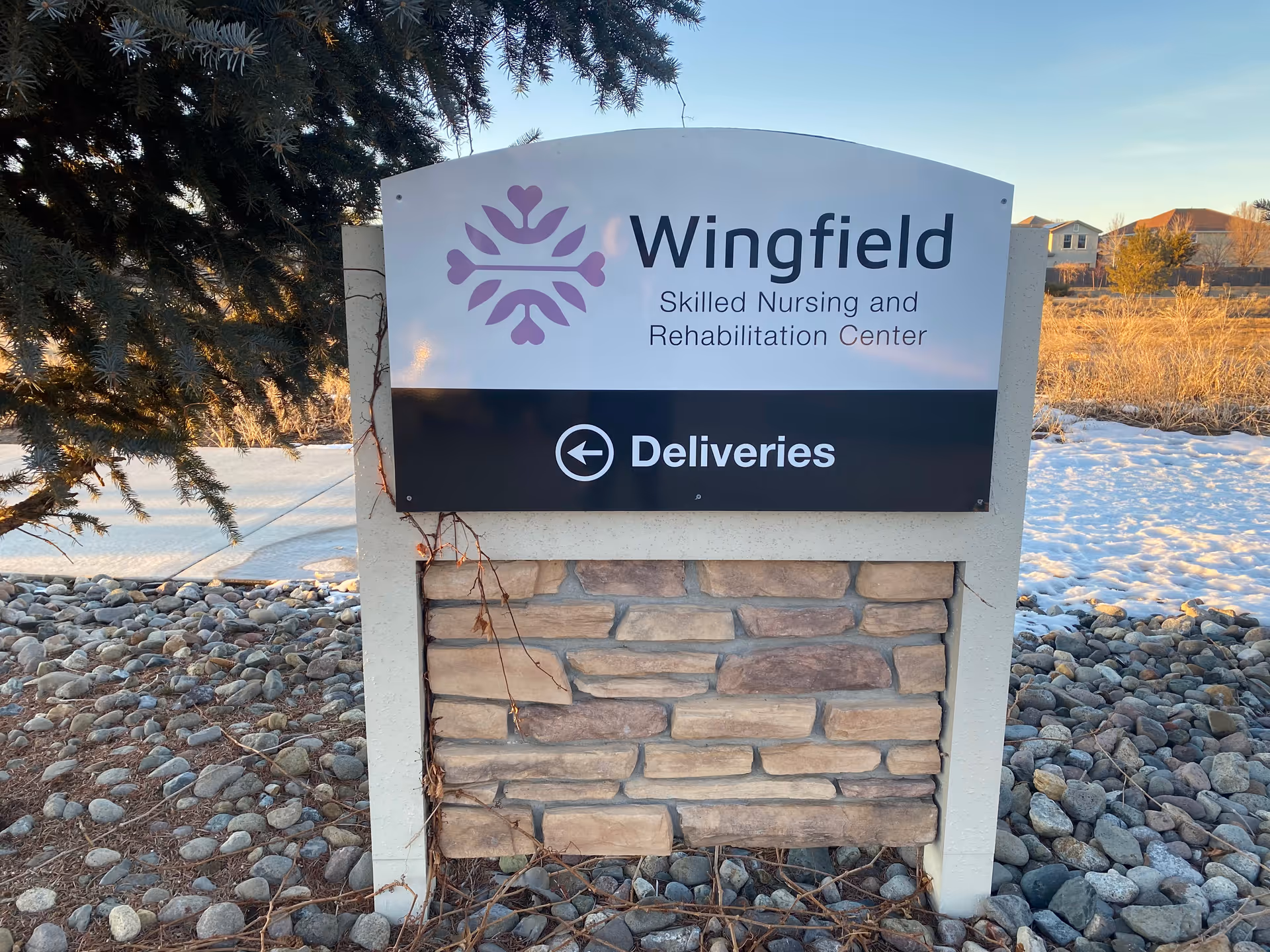 Outdoor sign for Wingfield Skilled Nursing and Rehabilitation Center with a delivery direction arrow, surrounded by rocks and some snow on the ground, with trees and houses in the background.