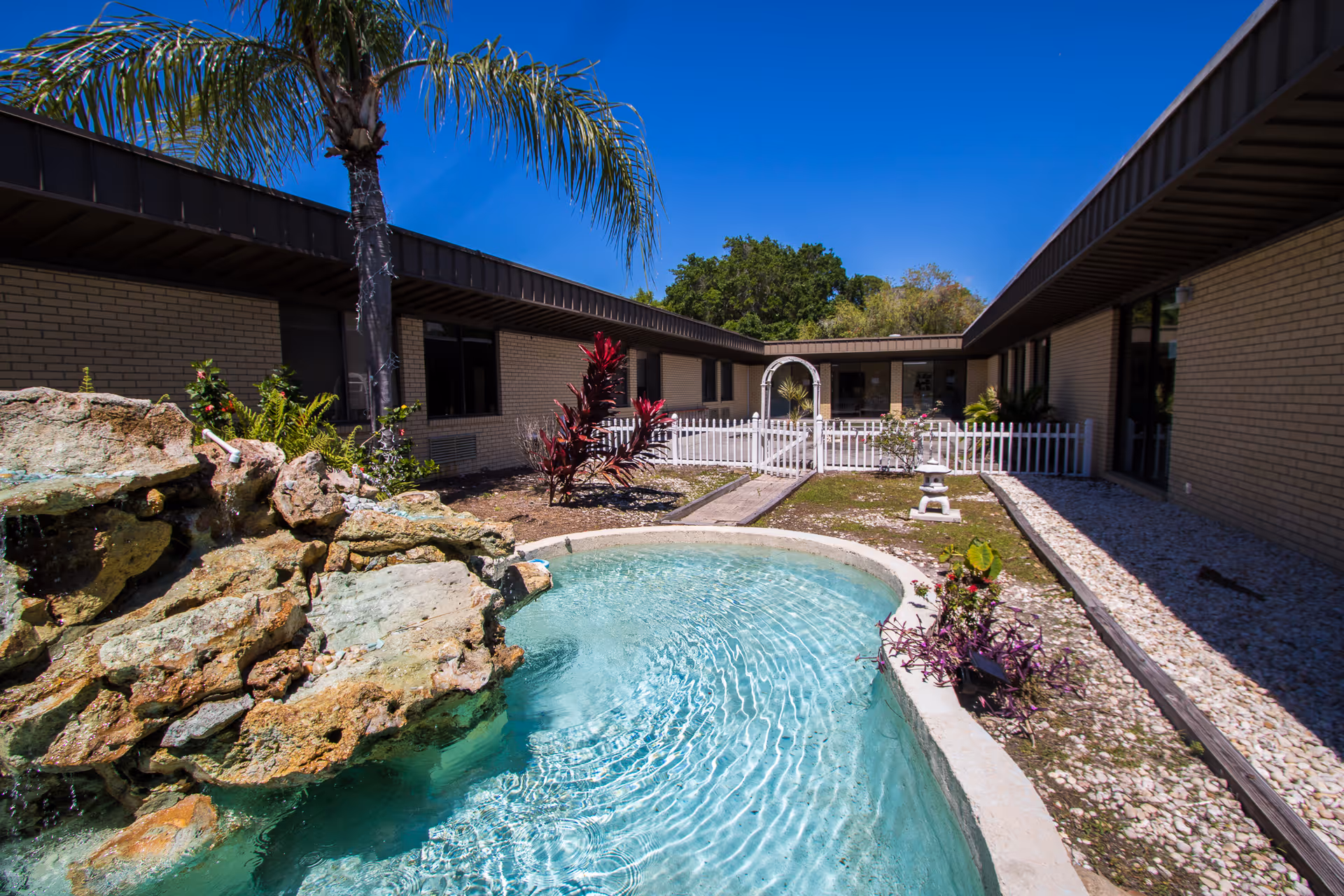 Sunny courtyard with a small turquoise pond and rock waterfall, palm tree, landscaping, a white picket fence and single-story brick building surrounding it.