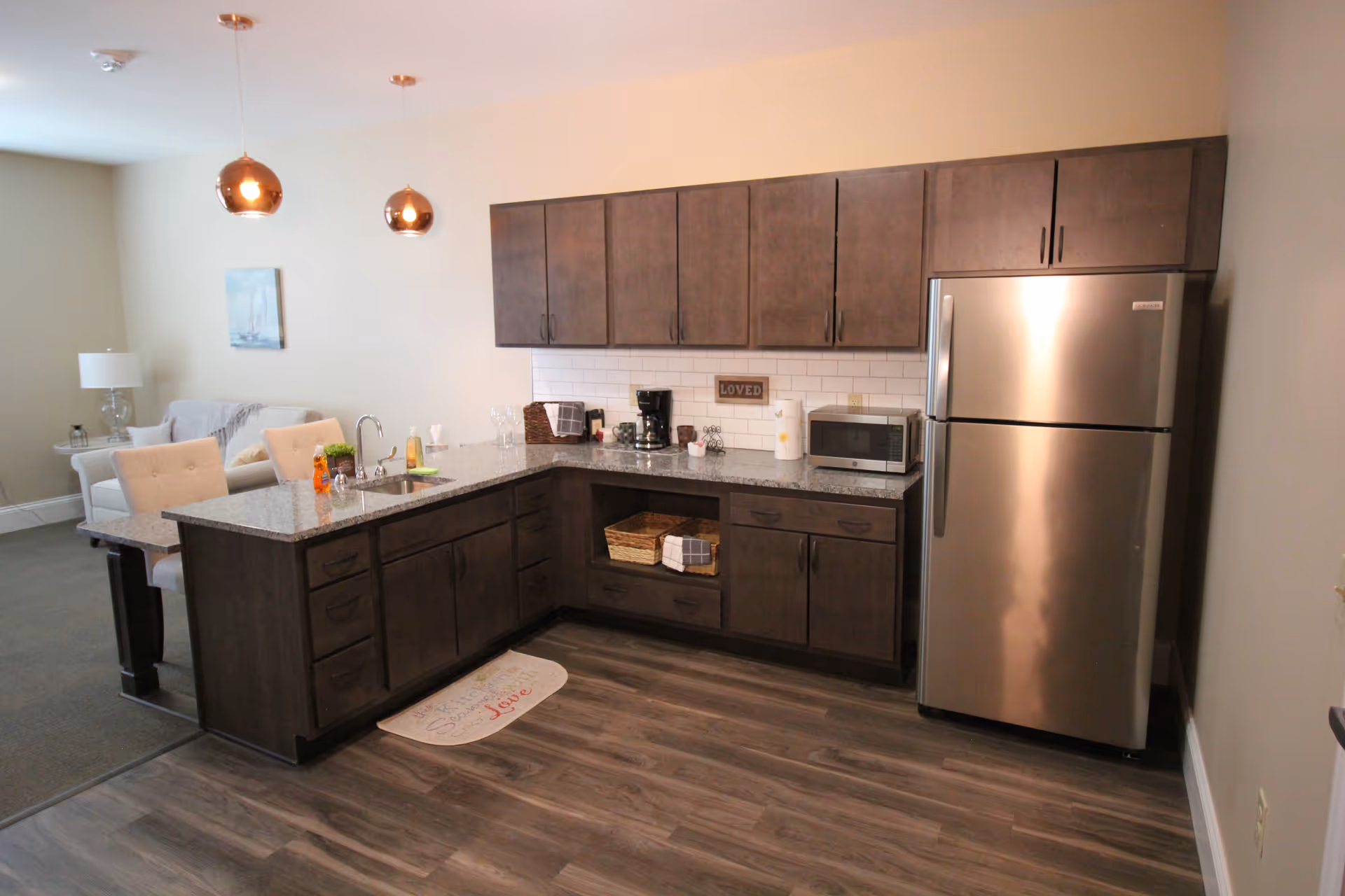 A modern kitchen area with dark wood cabinets, a granite countertop, a stainless steel refrigerator, a microwave, a coffee maker, and a sink. Two pendant lights hang from the ceiling above the counter, which has two beige upholstered chairs. In the background, there is a small living area with a light-colored armchair, a side table with a lamp, and a wall painting of a sailboat.