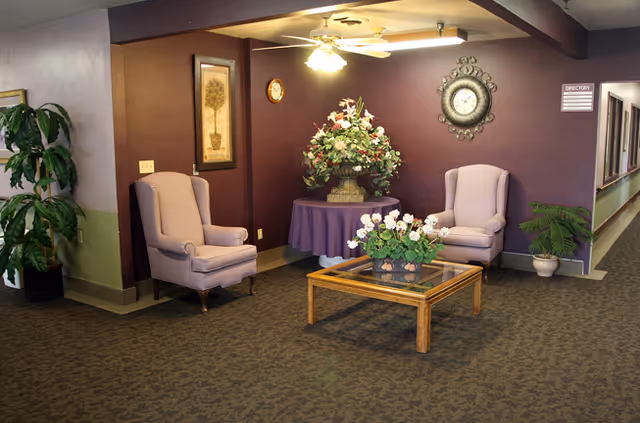 A cozy seating area in a senior living facility with two upholstered armchairs placed against a purple wall. Between the chairs is a round table covered with a purple tablecloth, holding a large floral arrangement. In front of the chairs is a wooden coffee table with a glass top and a smaller flower arrangement. The wall features a decorative clock and framed artwork. A ceiling fan with lights is above, and there are plants on either side of the seating area. A hallway extends to the right.