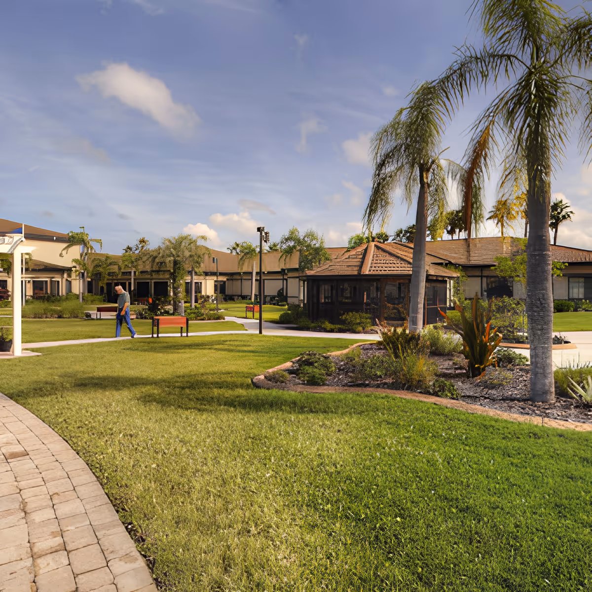 Grassy courtyard with palm trees, benches, and low buildings of a senior living community under a partly cloudy sky.
