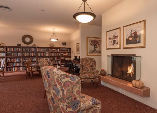 A cozy senior living common area with patterned armchairs facing a lit fireplace. The room features a carpeted floor, framed artwork above the fireplace, and bookshelves filled with books along the back wall. There are also computer desks with chairs in the background and ceiling light fixtures providing warm lighting.