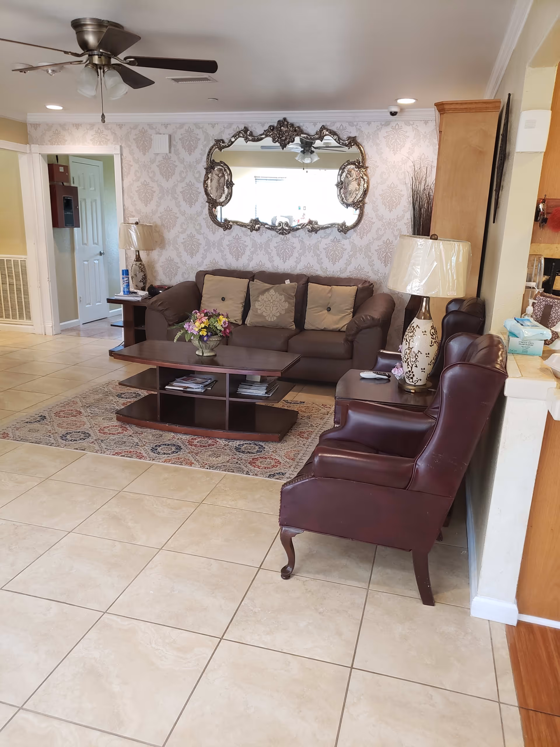 A cozy living room with a brown leather sofa adorned with beige and patterned cushions, a wooden coffee table with magazines underneath and a flower arrangement on top, two dark red leather armchairs, a decorative table lamp, and an ornate wall mirror reflecting a ceiling fan. The room has beige tiled flooring and patterned wallpaper.