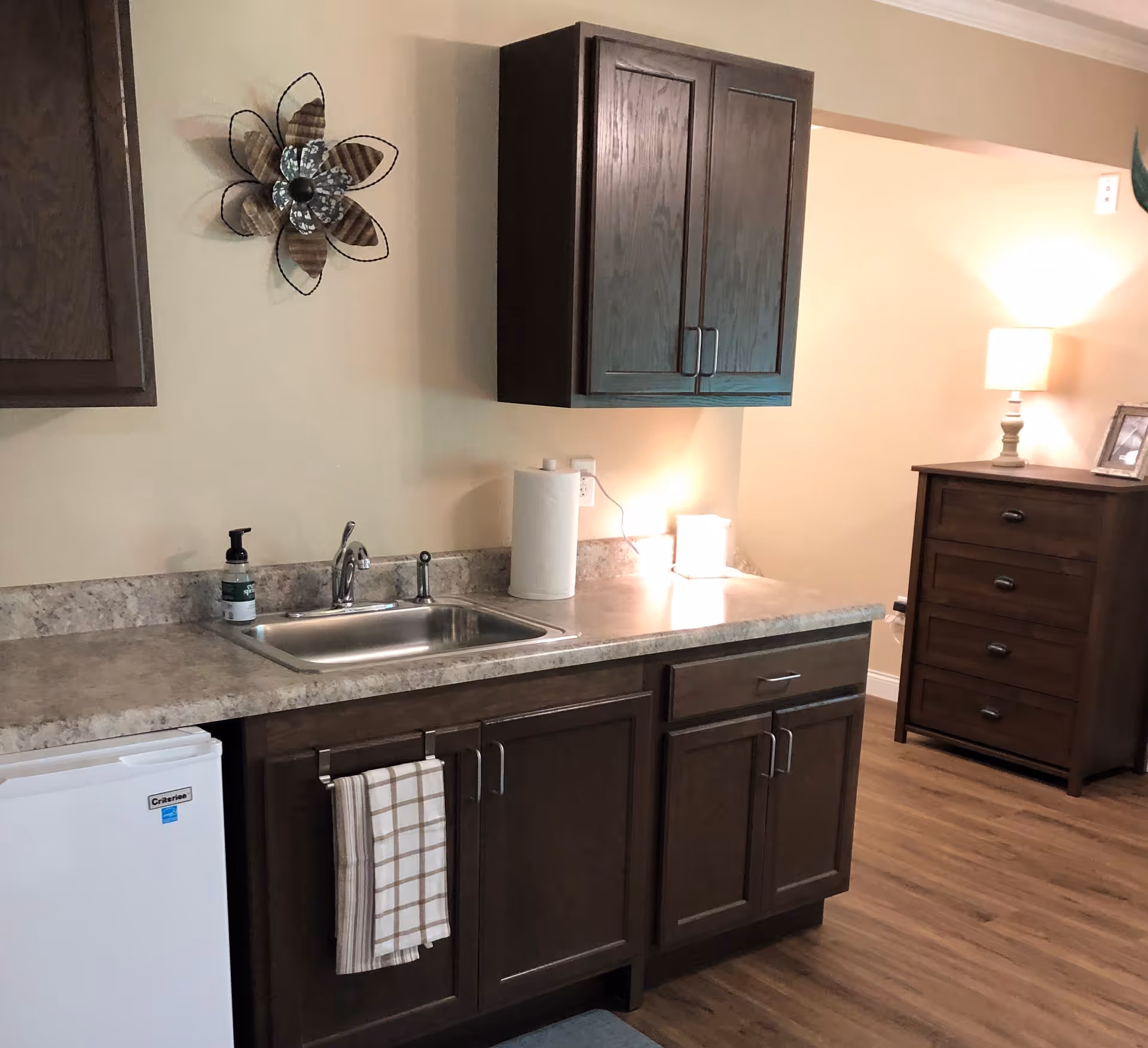 Interior view of a kitchenette area with a countertop, stainless steel sink, paper towel roll, soap dispenser, and dark wood cabinets above and below. A small white mini-fridge is visible under the counter. To the right, there is a wooden dresser with a lamp and a framed photo on top. The floor is wood, and the walls are painted beige. A decorative metal flower wall art is mounted above the sink.
