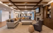 Spacious and well-lit living room area with modern furniture including sofas, armchairs, and a coffee table. The room features a stone fireplace with a mounted flat-screen TV above it, wooden ceiling beams, and built-in shelving units. The flooring is carpeted, and the overall decor is warm and inviting.