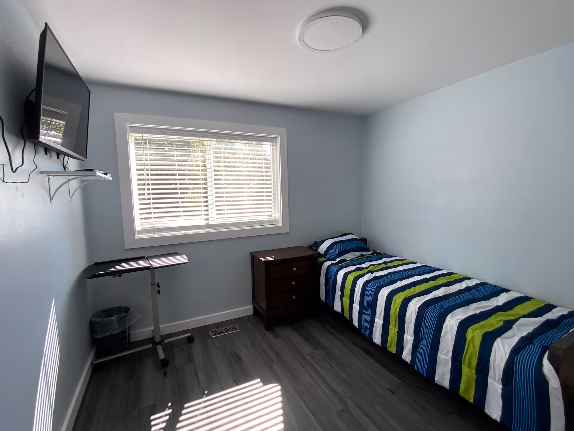 A small bedroom with light blue walls and a single bed with a striped blue, green, and white comforter. There is a wooden nightstand next to the bed, a window with white blinds letting in sunlight, a wall-mounted TV, a small rolling table, and a trash bin in the corner.