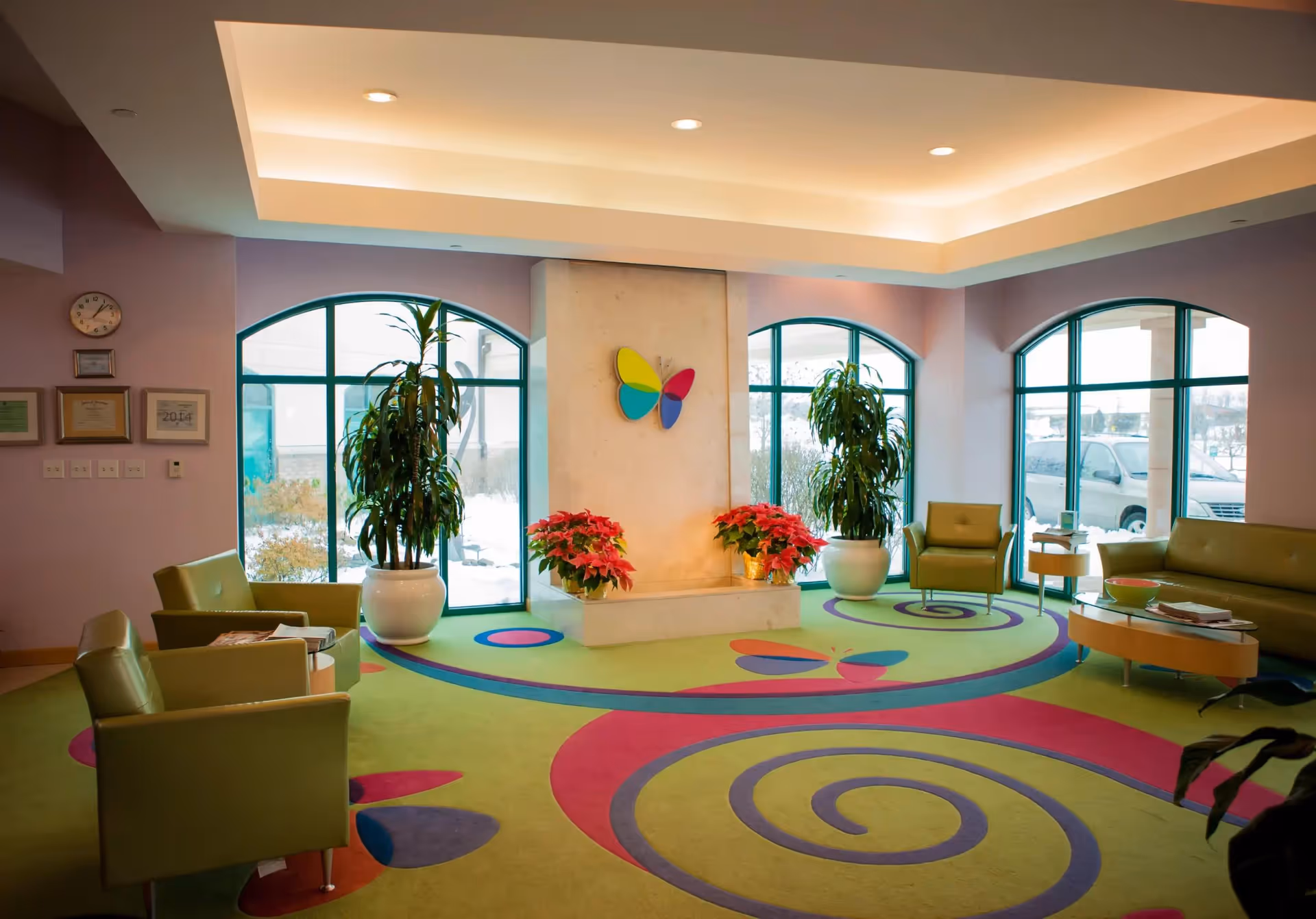 Bright, colorful lobby seating area with green chairs, patterned carpet, potted plants and a central wall featuring a butterfly decoration.
