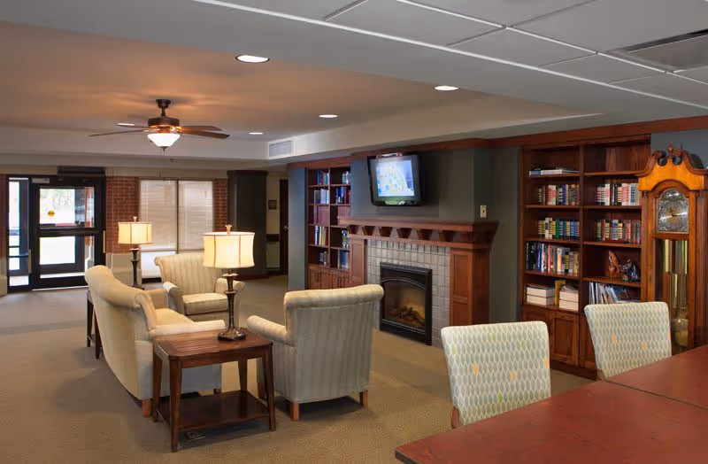 A cozy communal lounge with upholstered chairs and sofas facing a fireplace and wall-mounted TV, flanked by bookshelves and a grandfather clock.