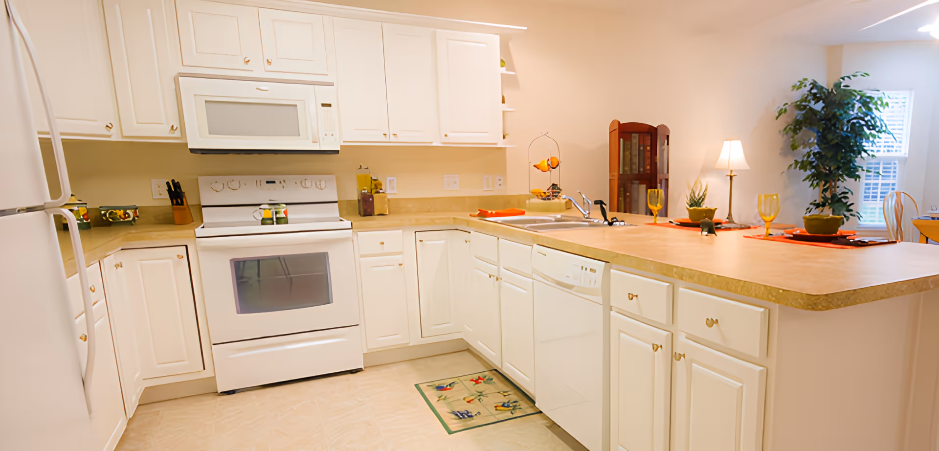Bright and clean kitchen with white cabinets, a white refrigerator, stove, microwave, and dishwasher. The kitchen has a beige countertop with a sink and faucet, and is decorated with a small plant, a fruit stand, and place settings with yellow glasses and orange placemats. There is a window with blinds and a tall green plant in the background.