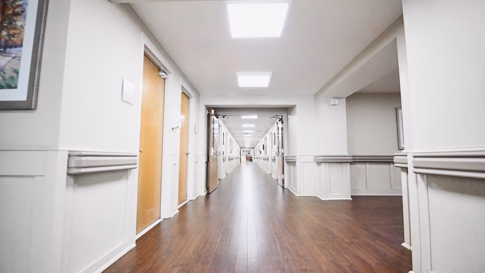 A long, well-lit hallway in a senior living facility with wooden floors, white walls, and multiple closed wooden doors on both sides. The ceiling has rectangular fluorescent lights, and there are handrails along the walls.