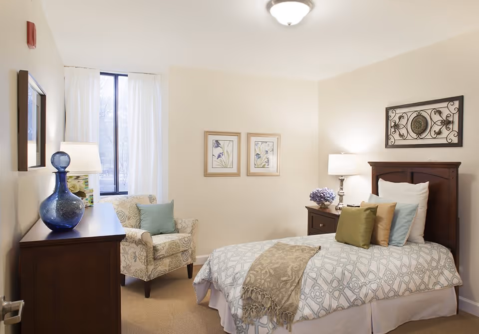 A cozy bedroom in a senior living facility featuring a single bed with multiple pillows and a patterned bedspread. There is a wooden nightstand with a lamp and a small flower arrangement beside the bed. A comfortable armchair with a cushion is placed near a window with white curtains. The walls are decorated with two framed floral prints and a decorative metal wall art piece above the bed.