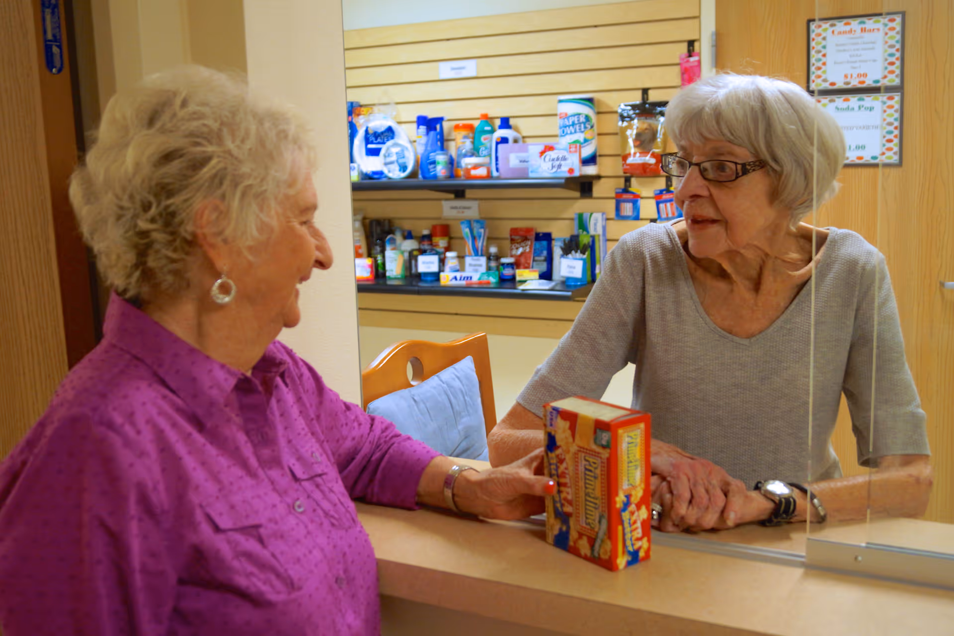 Two elderly women interacting at a counter in a small shop or reception area stocked with various household and personal care items. One woman in a purple shirt is standing on one side of the counter, while the other woman with gray hair and glasses is on the other side holding a box of crackers.