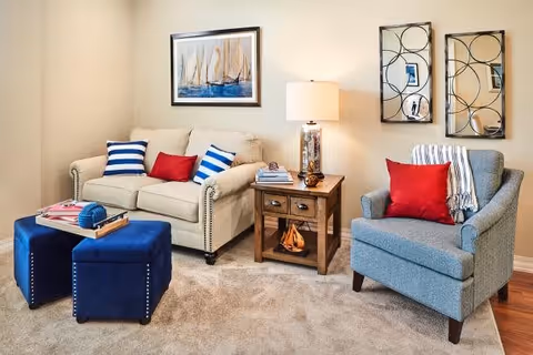 A cozy living room with a beige loveseat adorned with two blue and white striped pillows and one red pillow. Next to it is a wooden side table with a decorative lamp and books. A blue armchair with a red pillow and a striped throw blanket is positioned to the right. Two blue ottomans with nailhead trim are in front of the loveseat, holding a tray with a decorative item. The walls are decorated with a framed painting of sailboats and two decorative mirrors.