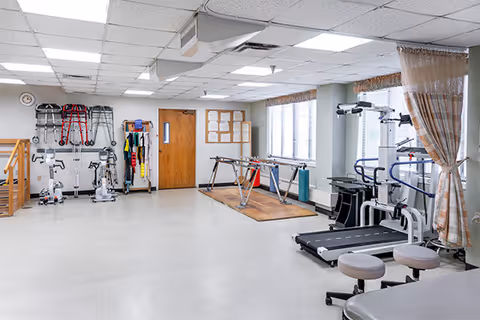 A spacious physical therapy or rehabilitation room with exercise equipment including treadmills, stationary bikes, parallel bars for walking practice, and various mobility aids such as walkers and canes hanging on the wall. The room has large windows with curtains, a wooden door, and a bulletin board on the wall.