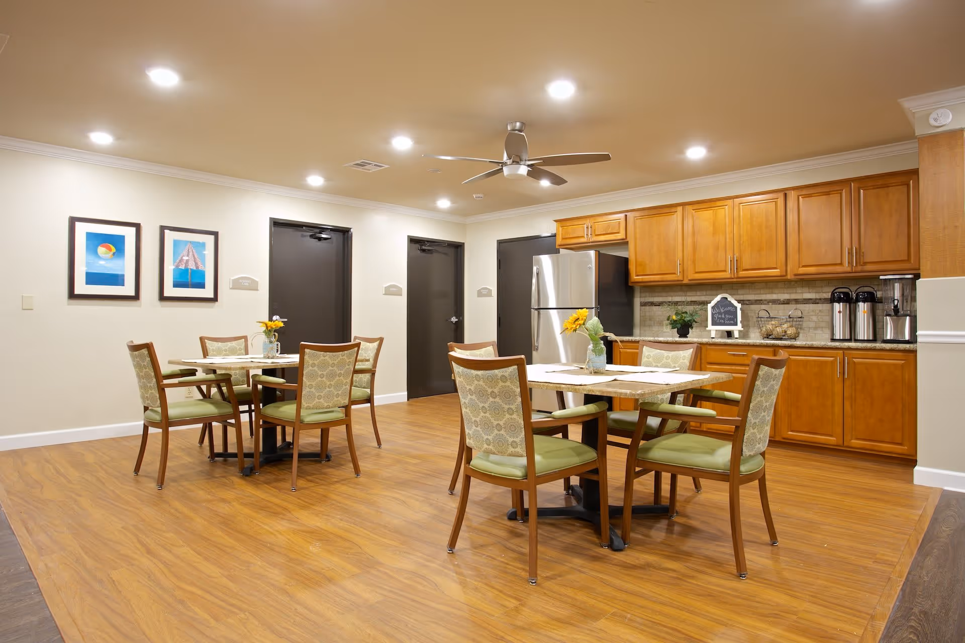 Well-lit dining area with round tables and chairs, wood flooring, and a kitchenette with wooden cabinets and a stainless refrigerator.