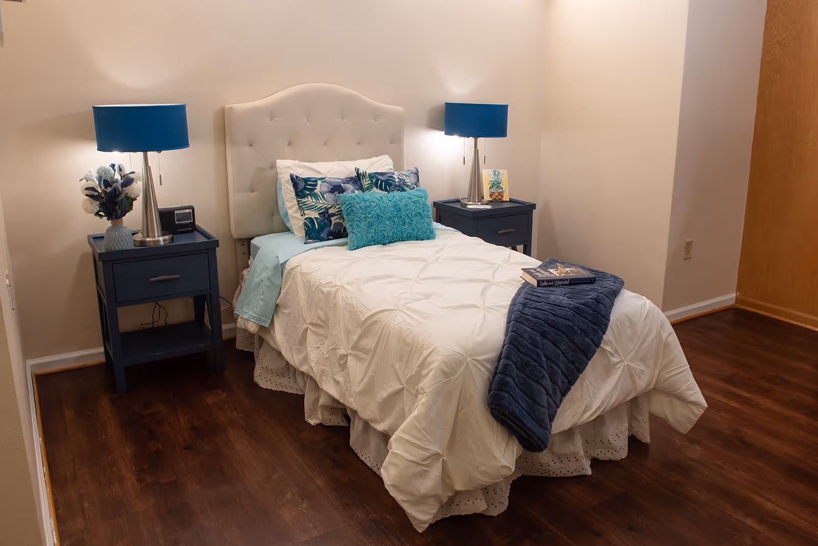 A neatly made single bed with a white tufted headboard, white comforter, and decorative pillows in shades of blue and green. There are two matching blue nightstands on either side of the bed, each with a blue lamp. One nightstand has a vase with flowers and a clock, while the other has a small framed picture. A dark blue throw blanket and a book are placed on the bed. The room has wooden flooring and beige walls.