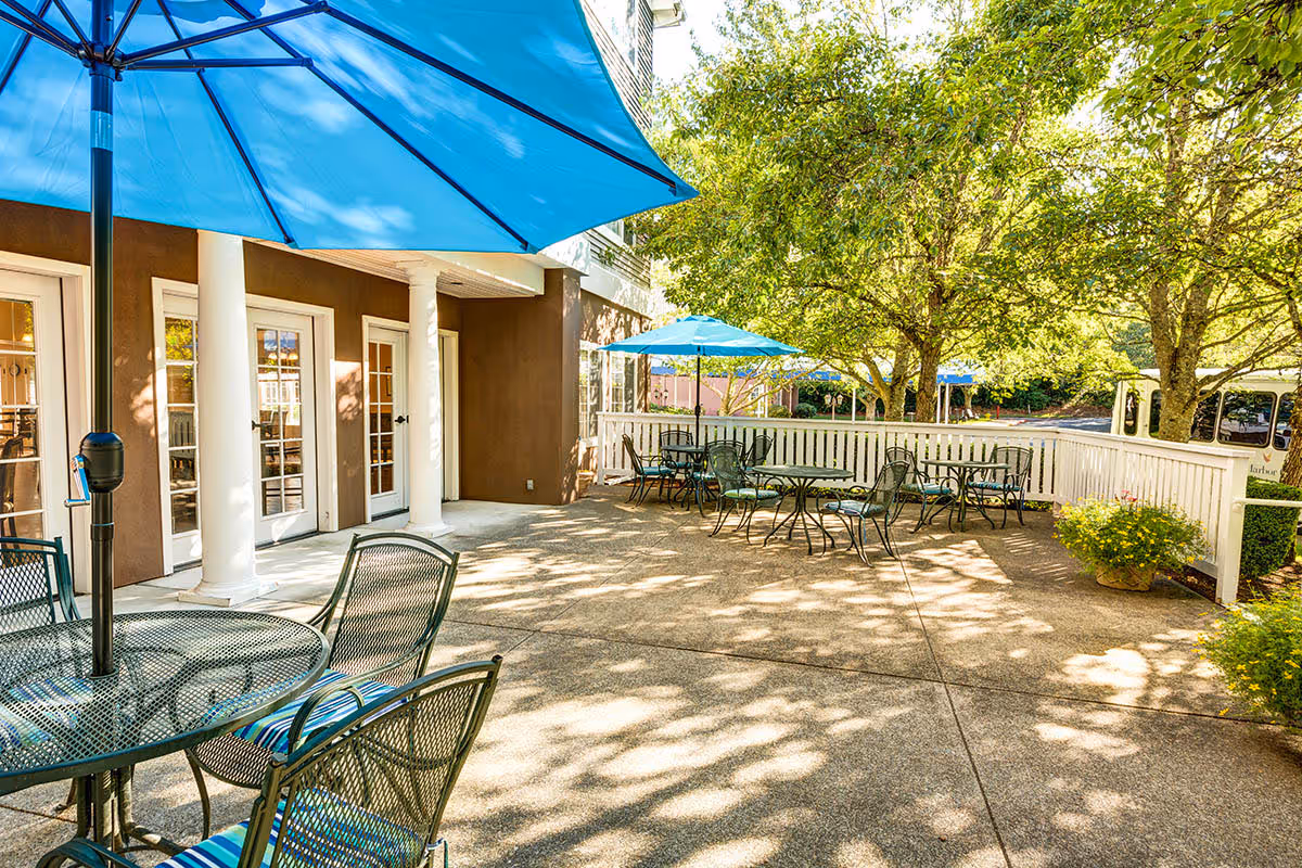 Outdoor patio area at a senior living facility with several metal tables and chairs, each shaded by large blue umbrellas. The patio is surrounded by a white fence and shaded by trees, with sunlight filtering through the leaves. The building exterior features brown walls and white columns with glass doors leading inside.