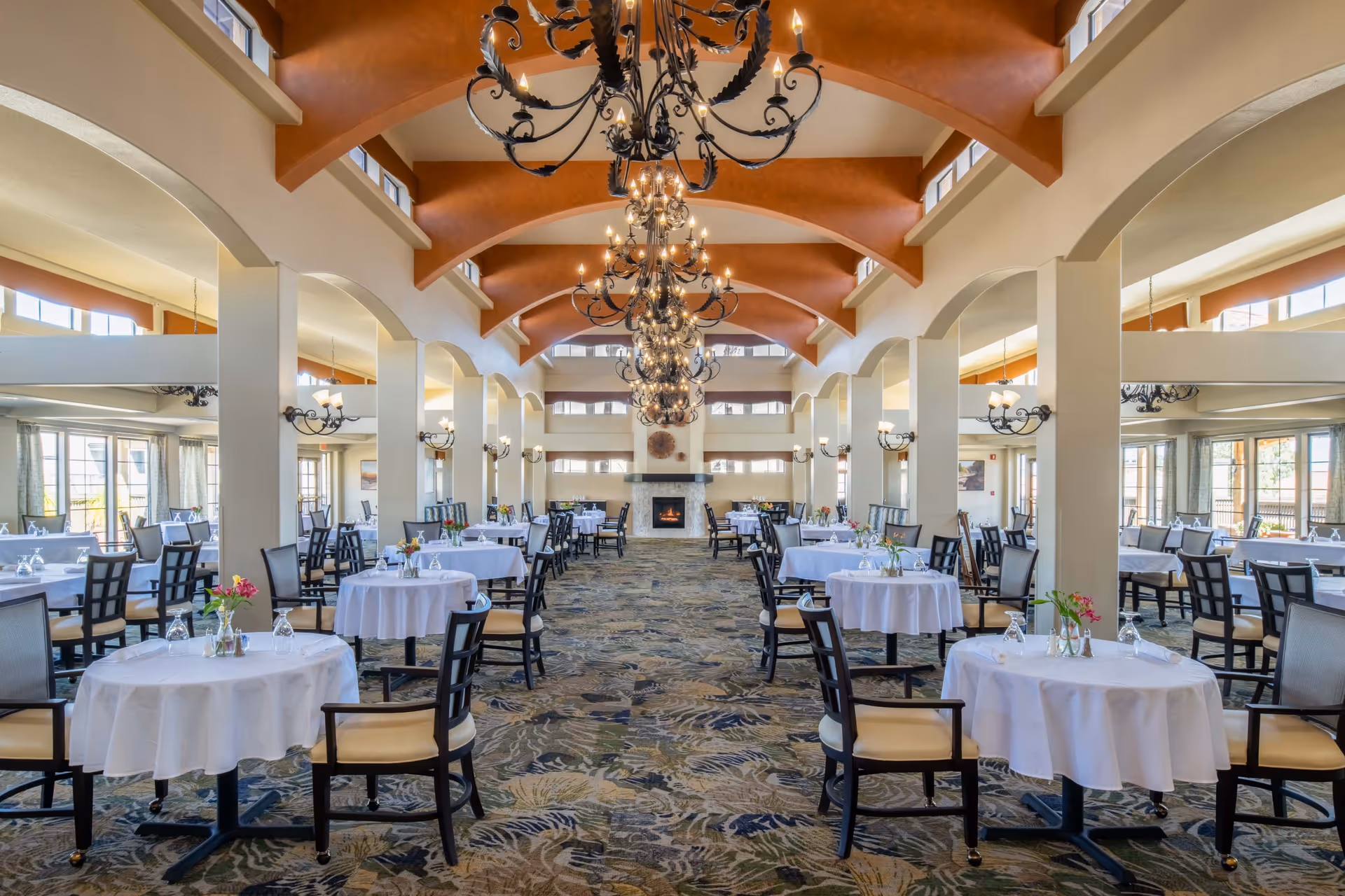 Spacious dining room with multiple round tables covered with white tablecloths, each set with glasses and small flower arrangements. The room features high vaulted ceilings with large chandeliers, tall windows allowing natural light, and a fireplace at the far end.