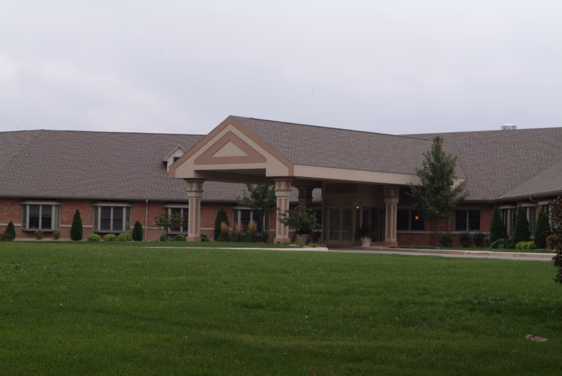 Front exterior view of a single-story brick building with a covered entrance supported by columns, surrounded by a well-maintained lawn and some small trees and shrubs.