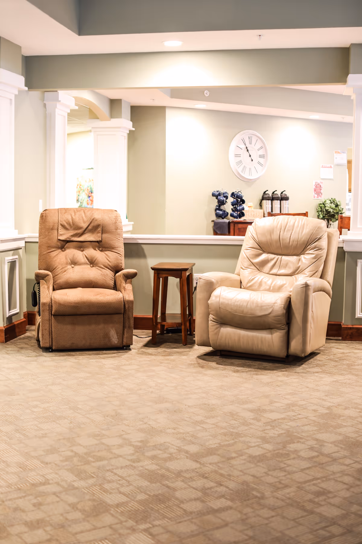Two recliner chairs, one brown fabric and one beige leather, positioned side by side with a small wooden side table between them in a well-lit room with light green walls. A large wall clock and some decorative items are visible in the background.
