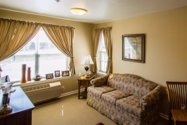 A cozy living room with beige walls and carpet, featuring a patterned sofa, a wooden side table with a lamp, framed pictures on the window sill, and two windows with beige curtains letting in natural light.