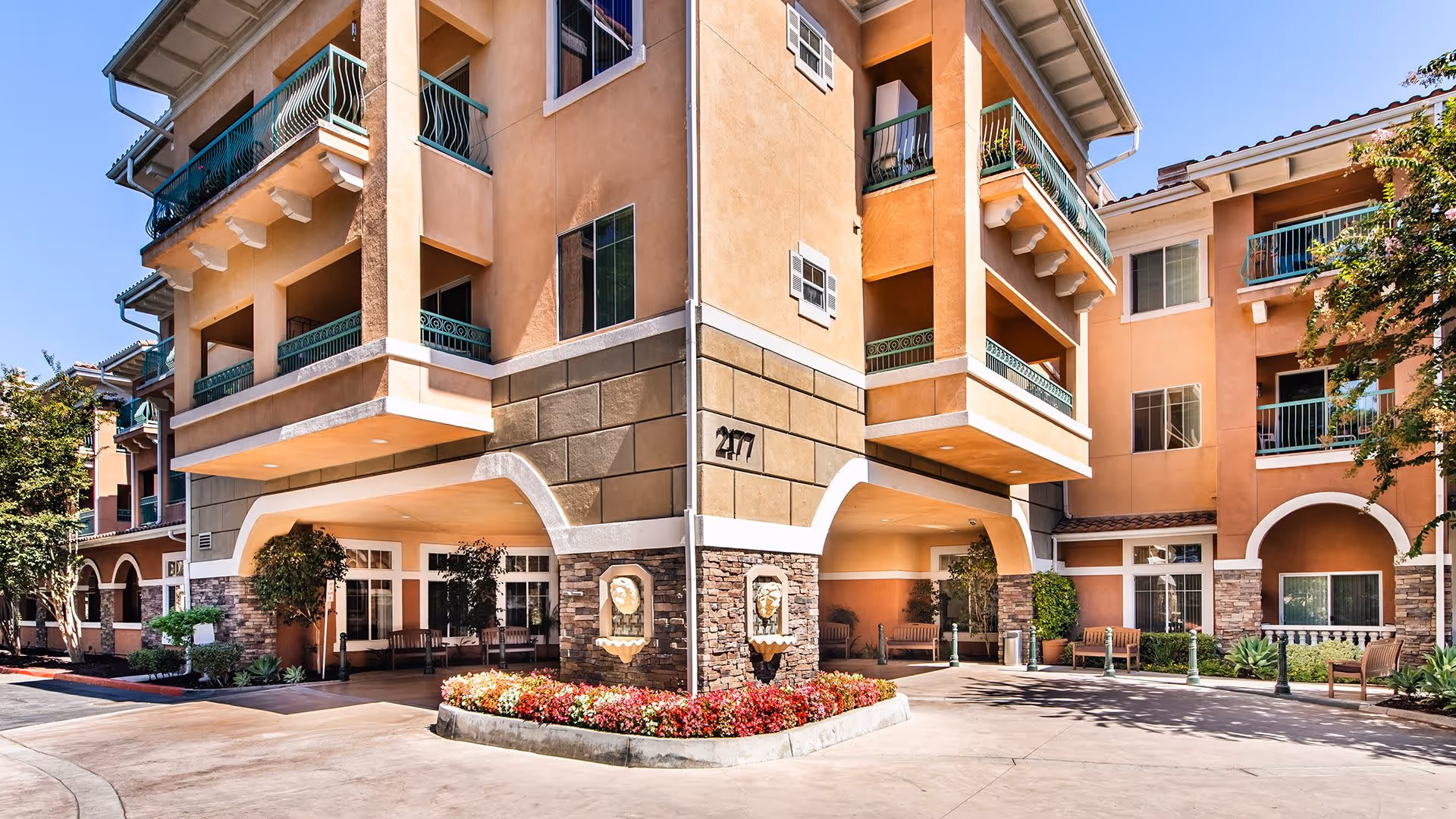 Exterior view of a multi-story senior living facility building with peach-colored walls, balconies with green railings, stone accents, and a landscaped flower bed at the entrance under a clear blue sky.