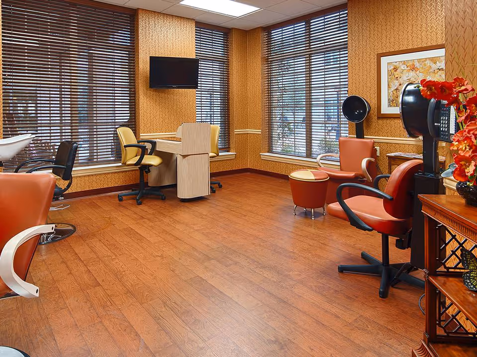 Interior of a salon room with wooden flooring, several salon chairs including red and black ones, a small desk with yellow chairs, large windows with wooden blinds, a wall-mounted TV, and a framed floral picture on the wall.