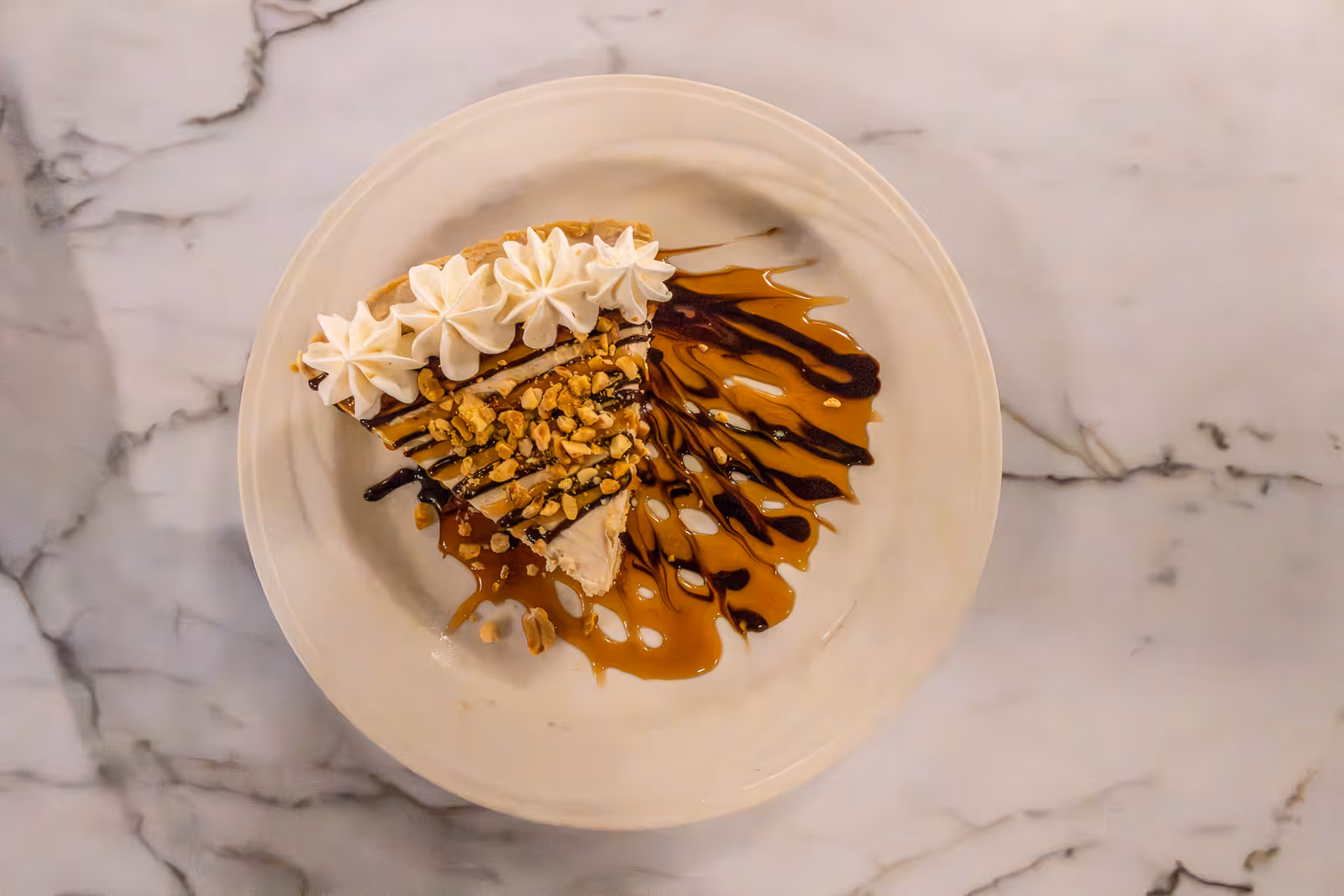 A slice of peanut butter pie topped with whipped cream and chopped peanuts, drizzled with chocolate and caramel sauce on a white plate, placed on a marble surface.