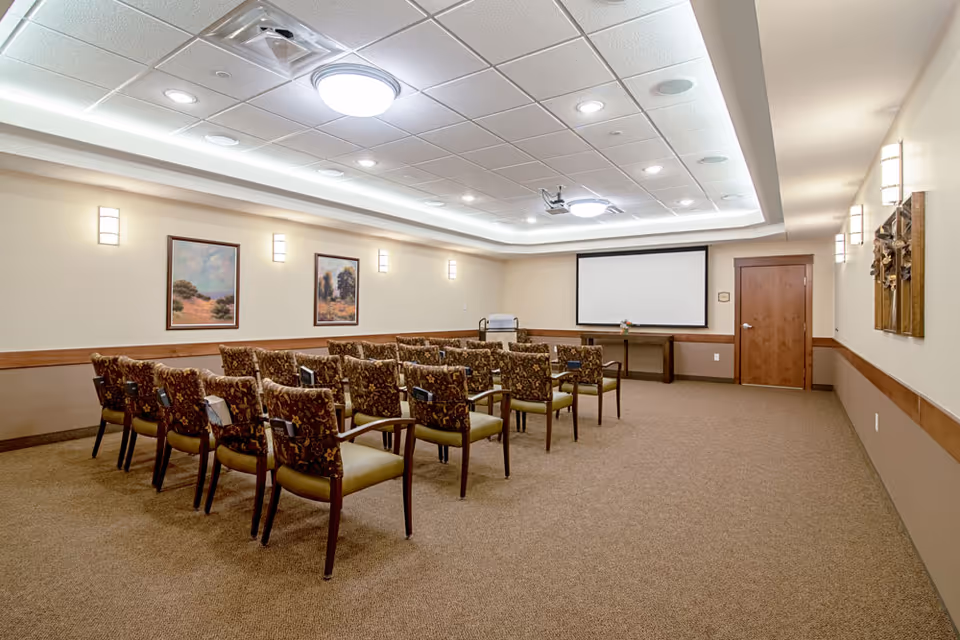 A small conference or presentation room with rows of patterned chairs facing a projector screen on the far wall. The room has beige walls, carpeted floor, framed landscape paintings, and ceiling lights.