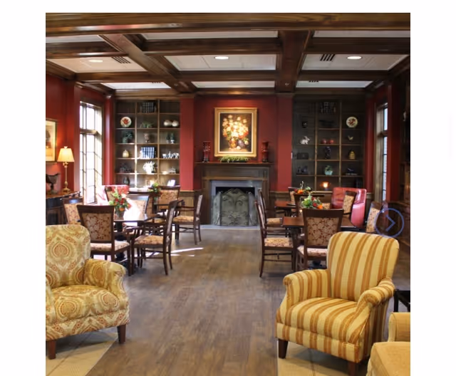 A cozy common area with wooden floors, multiple upholstered armchairs and dining tables with chairs. The room features a fireplace with a floral painting above it, built-in bookshelves on either side, and large windows letting in natural light. The walls are painted a warm red color, and there are decorative lamps and plants throughout the space.
