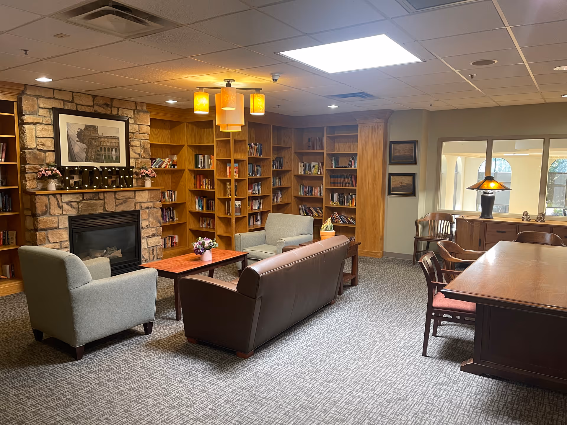 A cozy living room area with a stone fireplace, wooden bookshelves filled with books, a brown leather sofa, two gray armchairs, a wooden coffee table with a small flower arrangement, and a wooden dining table with chairs. Warm lighting fixtures hang from the ceiling, and there are framed pictures on the walls.