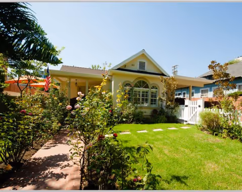 A single-story house with a well-maintained front yard featuring green grass, rose bushes, and a brick pathway leading to the front porch. The house has a light yellow exterior with a gabled roof and a large arched window. An American flag is displayed near the porch, and neighboring houses are visible in the background under a clear blue sky.