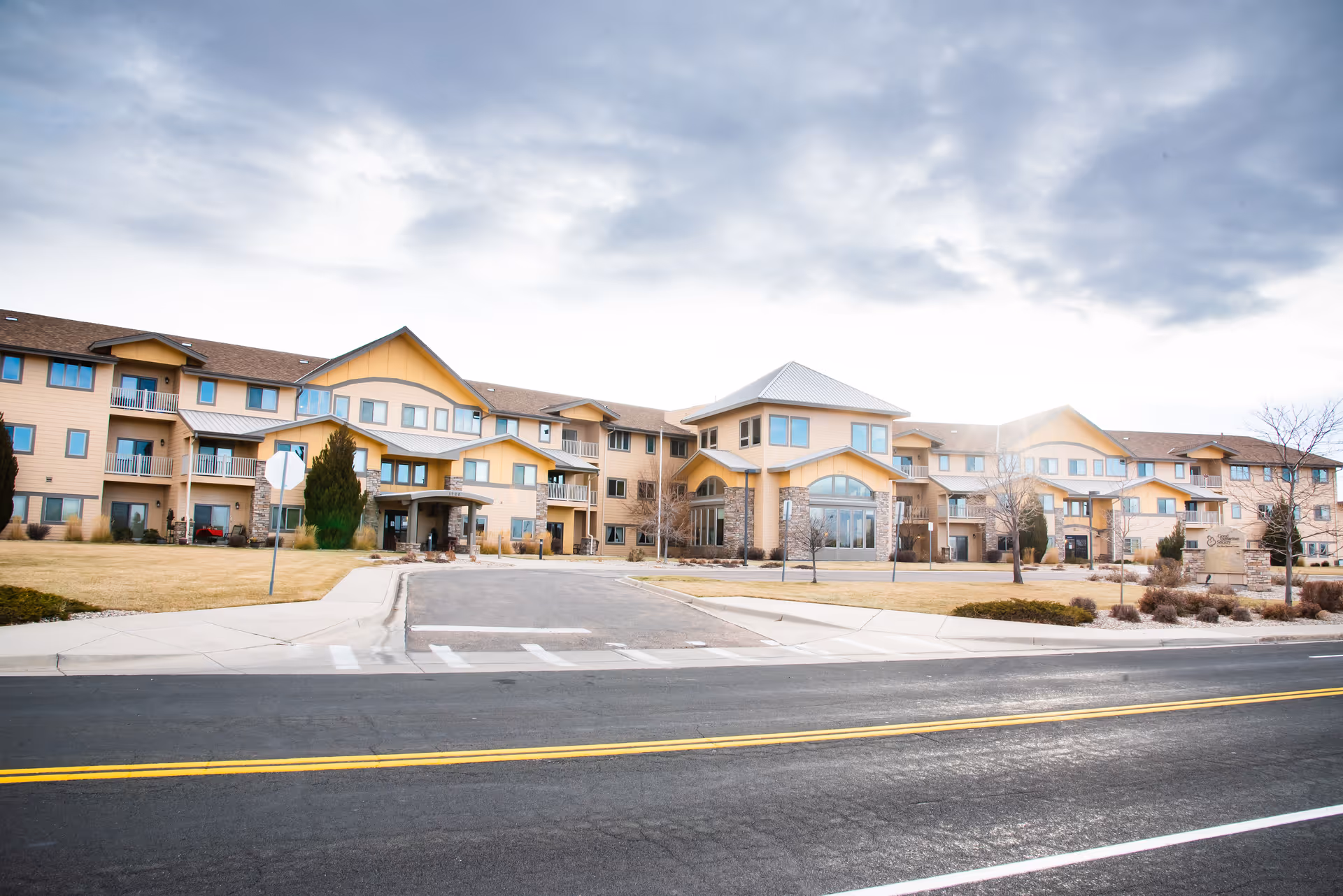 Wide frontal view of a multi-story senior living facility with a driveway, lawn, and cloudy sky.