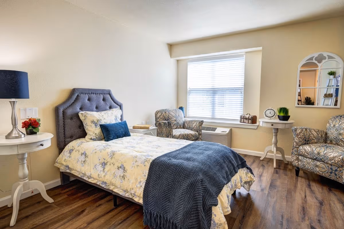 A cozy bedroom with a single bed featuring a tufted gray headboard, floral bedding, and a blue throw blanket. There are two patterned armchairs near a window with blinds, two white round side tables with decorative items including a lamp, flowers, a clock, and a small plant. A decorative mirror hangs on the wall, and the room has wooden flooring.
