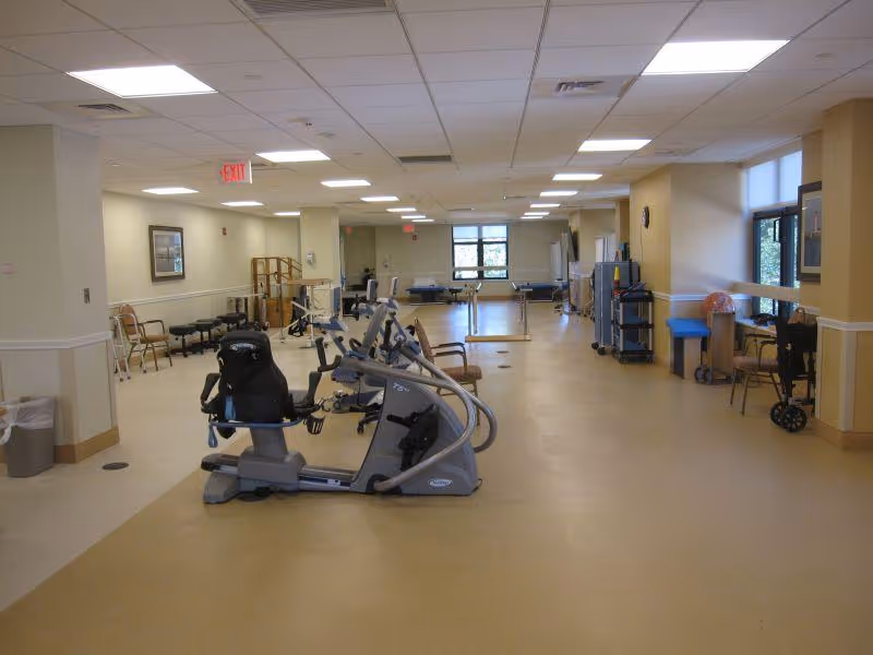 A spacious rehabilitation room with exercise equipment including stationary bikes and parallel bars. The room has beige flooring, cream-colored walls, fluorescent ceiling lights, chairs along the walls, and windows letting in natural light.