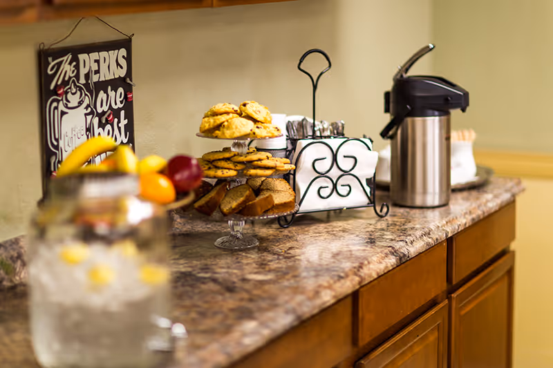 A countertop with a tiered tray holding cookies and bread slices, a metal napkin holder with white napkins, a stainless steel coffee dispenser, and a glass container with ice water and lemon slices. A decorative sign on the wall reads 'The Perks are Best'.