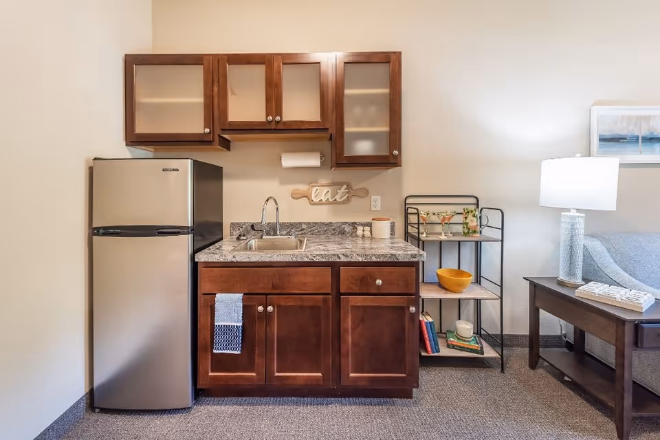 Small kitchenette area with a stainless steel refrigerator, dark wood cabinets with frosted glass doors, a granite countertop with a sink, and a towel hanging on the cabinet door. To the right, there is a small shelving unit with decorative items and books, and a side table with a lamp and a couch partially visible.