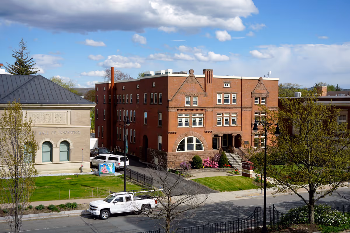 Exterior view of a red brick building with multiple windows and a stone base, surrounded by green lawns and trees under a partly cloudy sky. A white pickup truck is parked on the street in front of the building. Another building with the inscription 'Hall of Innovation' is visible to the left.