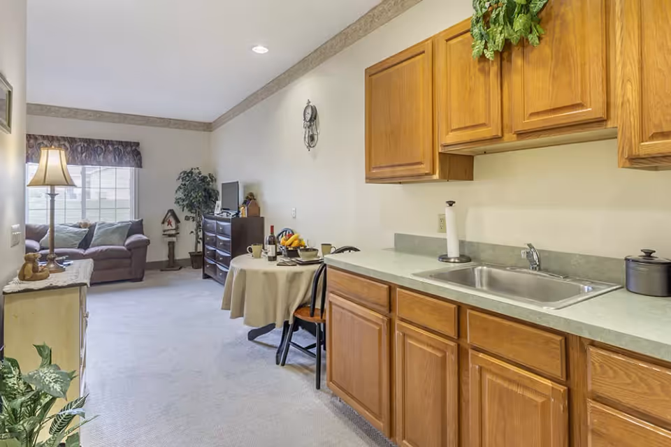 Interior view of a senior living facility room featuring a small kitchen area with wooden cabinets, a countertop with a sink, and a paper towel holder. Adjacent to the kitchen is a round table with two chairs, set with a bottle of wine, fruit bowl, and cups. In the background, there is a living area with a brown sofa, a floor lamp, a TV on a dresser, and a potted plant near a window with a valance.