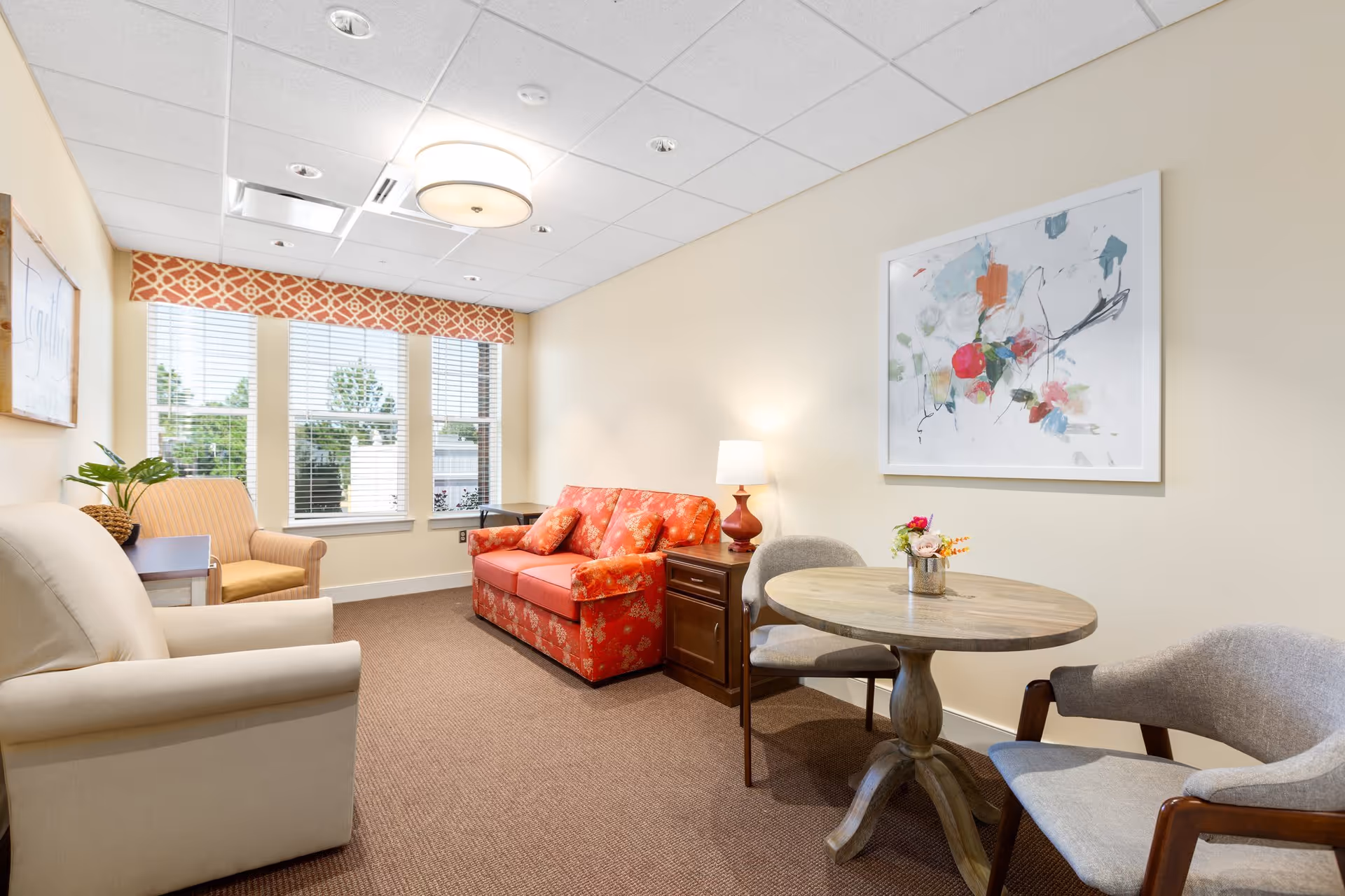 A cozy living room with a beige armchair, a striped armchair, and a red floral sofa. There is a wooden side table with a lamp next to the sofa, and a round wooden table with two gray upholstered chairs. A colorful abstract painting hangs on the wall, and three large windows with blinds and a patterned valance let in natural light.